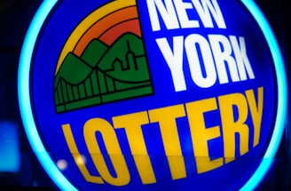 A bright, circular neon sign with a blue background, featuring the words 'New York Lottery' in bold white and yellow letters. The sign includes a colorful illustration of a landscape with green mountains, an orange-red sunset, and a bridge, all encapsulated within the circle.