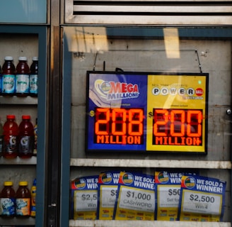 An advertisement display in a store window features lottery jackpot amounts for Mega Millions and Powerball, both showing jackpots of 208 million and 200 million respectively. Below the display, several lottery tickets and promotional posters are visible. To the left of the display, there is a refrigerator stocked with various drinks including different flavors of sports drinks and bottled water.