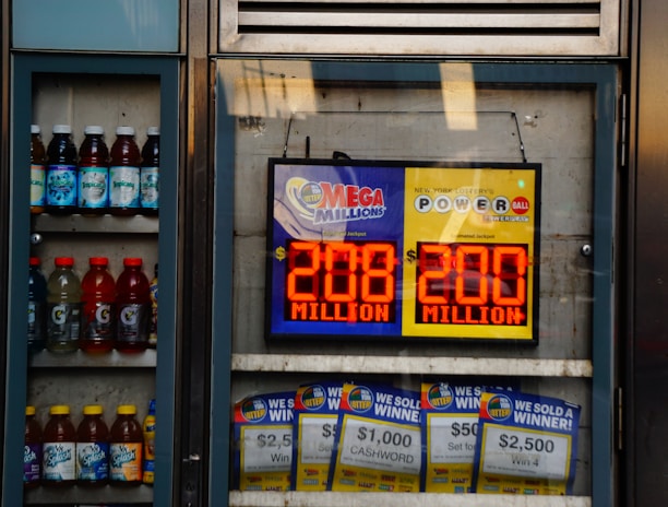 An advertisement display in a store window features lottery jackpot amounts for Mega Millions and Powerball, both showing jackpots of 208 million and 200 million respectively. Below the display, several lottery tickets and promotional posters are visible. To the left of the display, there is a refrigerator stocked with various drinks including different flavors of sports drinks and bottled water.