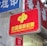 A red sign displaying the text 'China Welfare Lottery' in both Chinese and English, mounted on the exterior of a building. The surroundings include a blue sign with Chinese characters and a partial tree trunk visible on the right side.