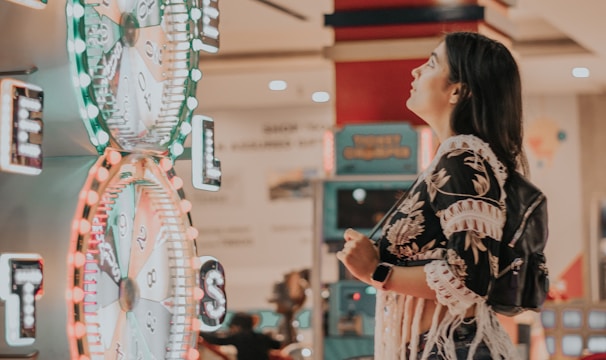 A woman is standing in an arcade looking at a large colorful game machine with bright lights. The game machine features large spinning wheels labeled with 'tickets' and has a vibrant design. The woman is casually dressed in a jacket with fringe details and is holding something in her hand while wearing a smartwatch.