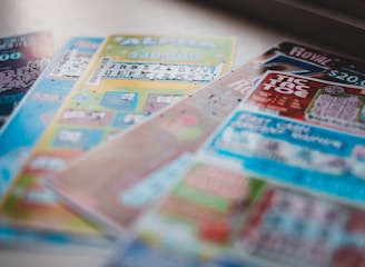 A selection of colorful lottery scratch-off tickets placed on a flat surface near a window. The image has a shallow depth of field, focusing on the tickets while blurring the background. The tickets display various games and prize amounts.