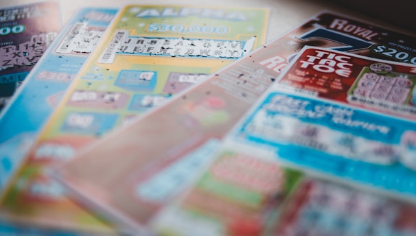 A selection of colorful lottery scratch-off tickets placed on a flat surface near a window. The image has a shallow depth of field, focusing on the tickets while blurring the background. The tickets display various games and prize amounts.