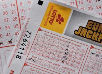 A close-up image of lottery tickets featuring crossed-out numbers, brand logos, and various numbers in a grid format. The tickets have sections for different games, and numbers are marked with black crosses.