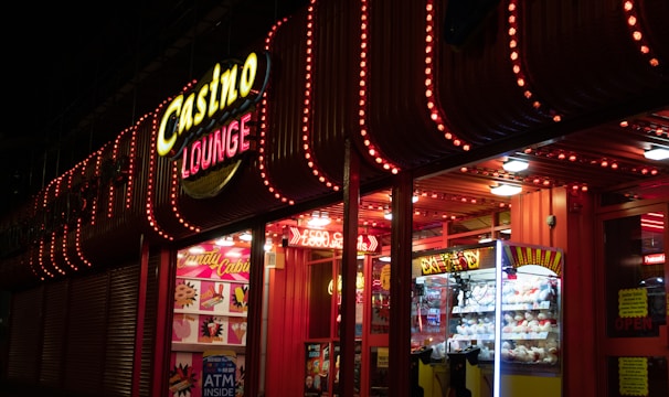 A brightly lit casino lounge entrance adorned with neon lights. The exterior features colorful signage including the words 'Casino' and 'Lounge' in vivid colors, with additional lights framing the building. Inside, a glimpse of a claw machine filled with plush toys and arcade games is visible. The area exudes an inviting yet bustling atmosphere typical of entertainment venues.