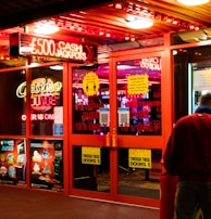 A brightly lit entrance to a casino with neon signs, including one advertising a £500 jackpot. The doors have clear signage indicating entry is restricted to those over 18. A person in a red jacket stands to the right, possibly interacting with a machine or display.
