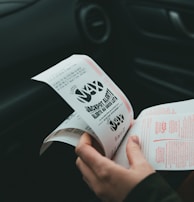 A person is holding and examining what appears to be lottery tickets inside a car. The image is focused on the tickets and the person's hands, with parts of the car interior visible in the background.