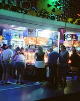 A group of people standing in front of an illuminated amusement arcade, engaging with various gaming machines. The environment is bustling with activity, and colorful lights create a vibrant atmosphere. The ceiling is decorated with large, playful images of dice and other game-related graphics, enhancing the lively mood.