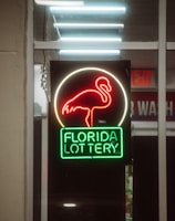 A neon sign depicting a flamingo above the words 'Florida Lottery' is prominently displayed. The sign is illuminated in bright red and green colors, and it is framed by glass panels. An 'EXIT' sign is visible in the background.