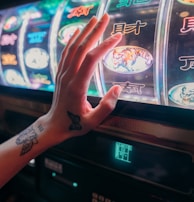 A hand with tattoos reaches towards a brightly lit slot machine, featuring various colorful symbols including a tiger. The image captures the moment of anticipation as the person engages with the machine during a game of chance.