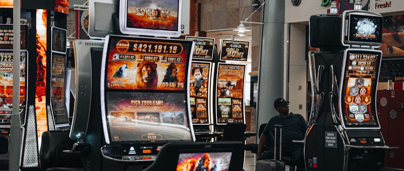 A collection of slot machines is situated inside a building with bright, colorful screens displaying various games and gaming themes. A person is seated nearby, seemingly relaxed with luggage beside them, and the atmosphere is lively with electronic displays.